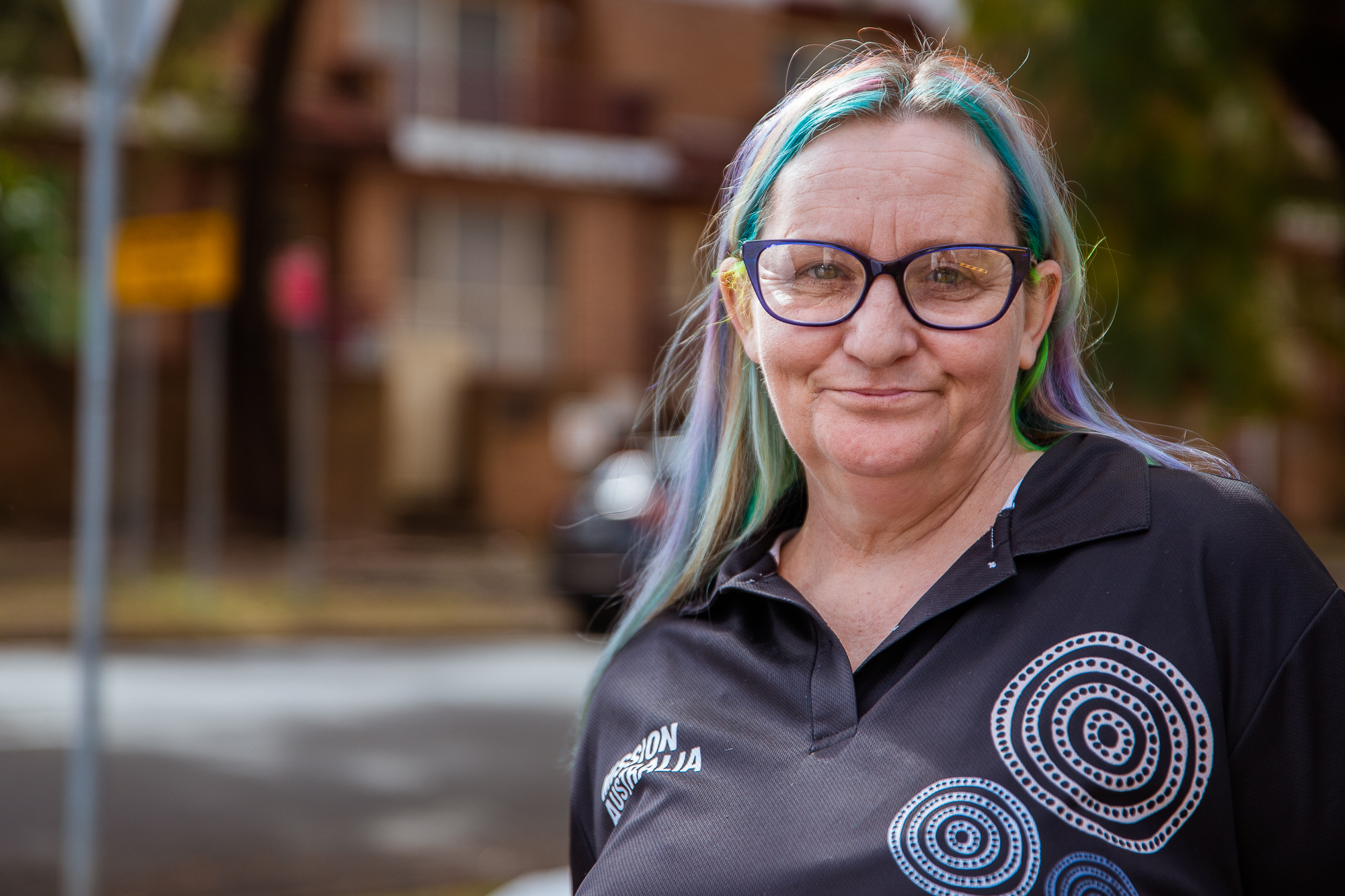 Kassie Paice standing outside in front of houses, with colourful hair and wearing a Mission Australia shirt.