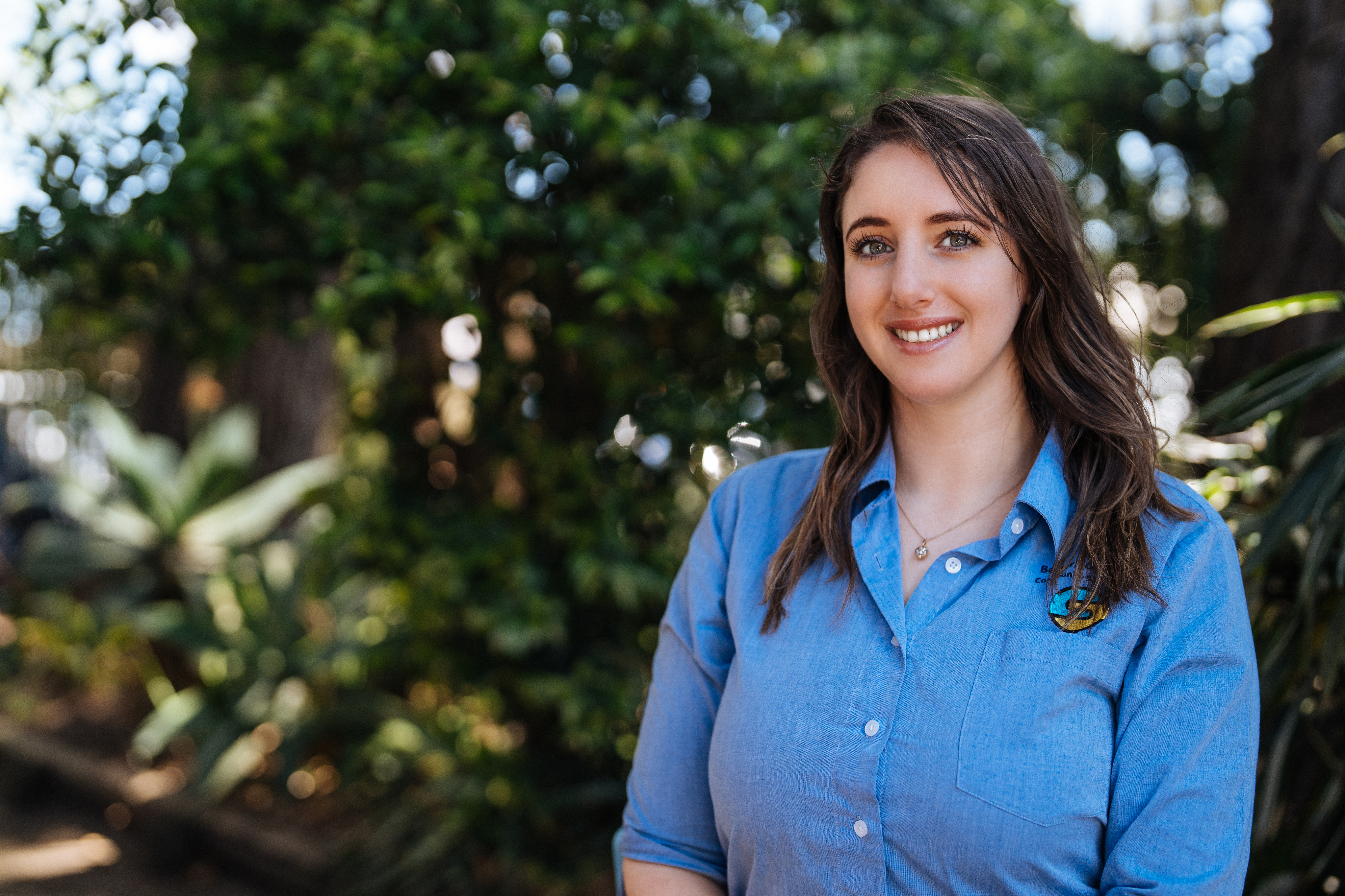 Riham Zeini wearing a blue shirt with trees behind her, standing and smiling at the camera.
